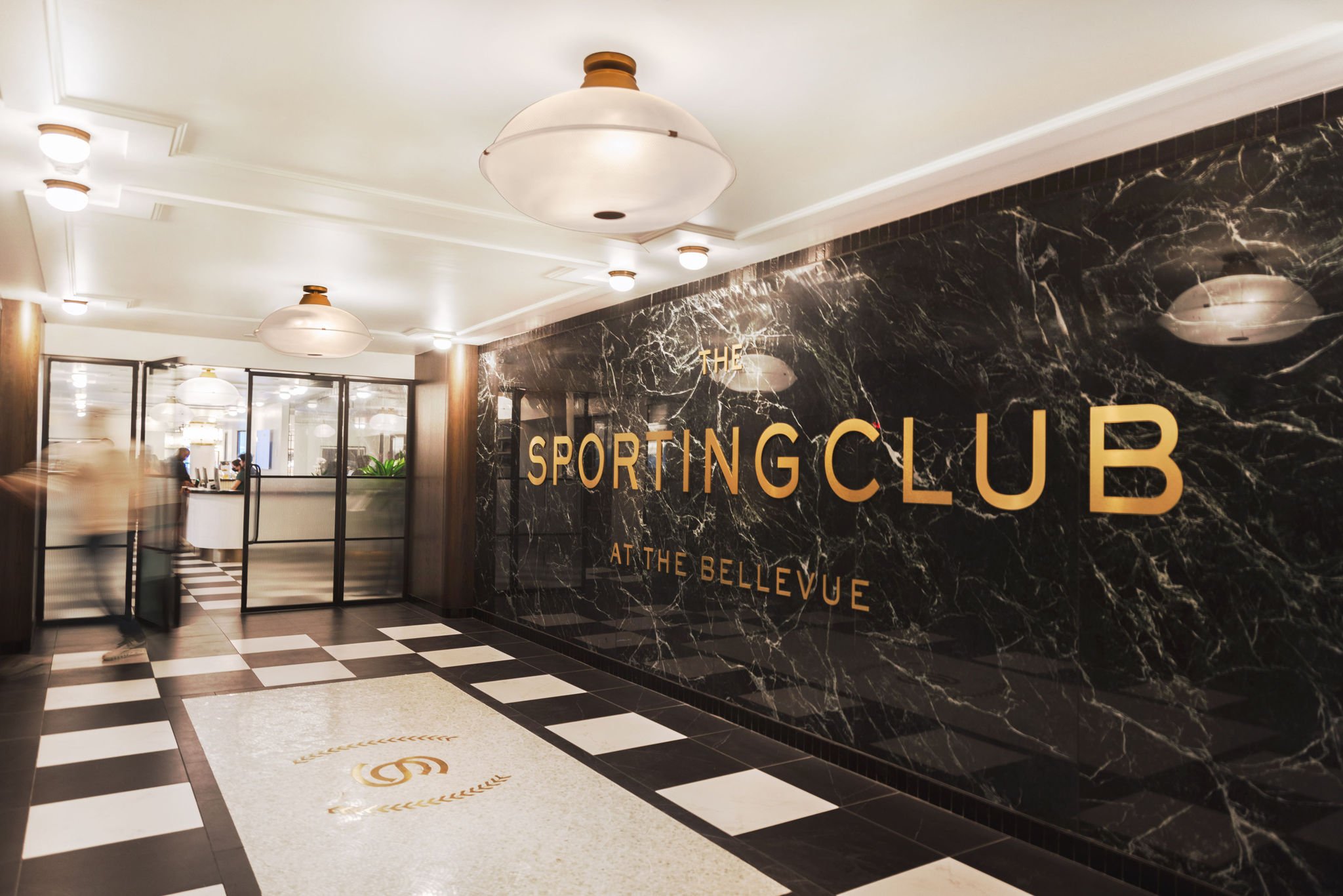 Entrance to the sporting club at The Bellevue. A marble wall displays the name next to black and white tiled floor
