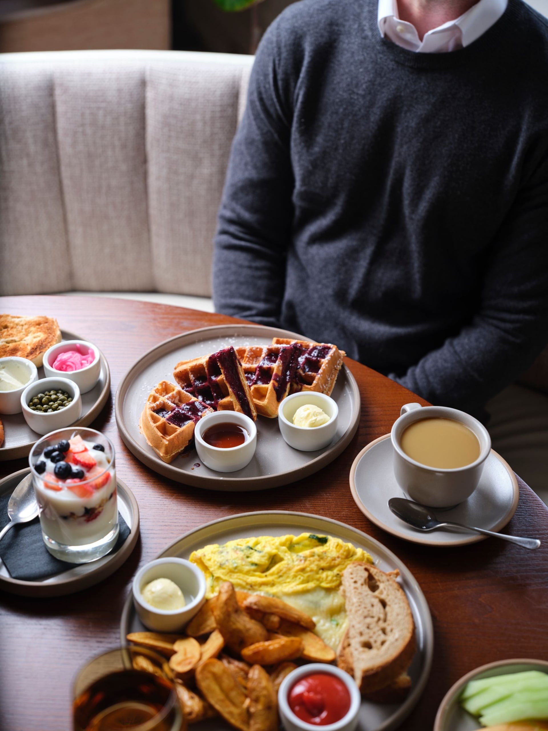 Person in a dark sweater sits at a wooden table with waffles, berry toppings, small bowls of condiments, and a cup of coffee nearby.
