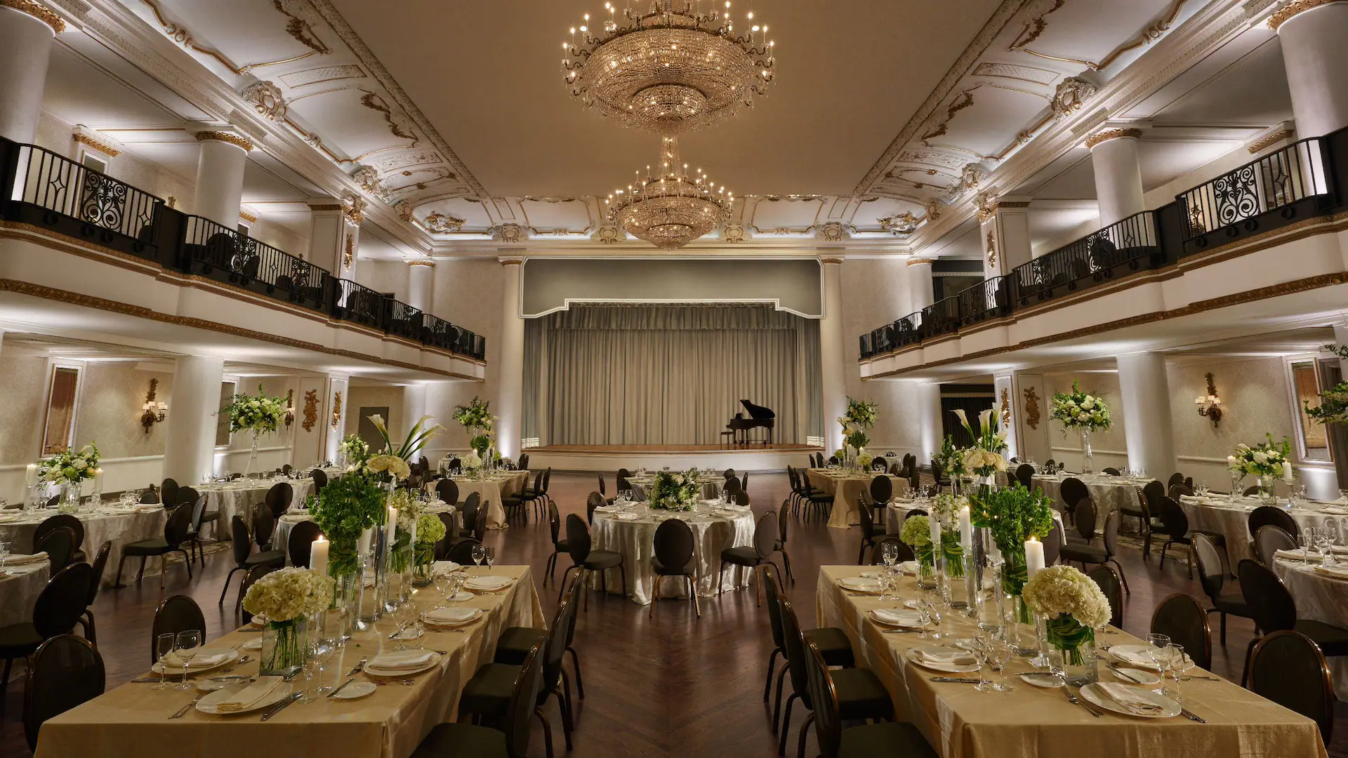 A wide shot of the Grande Belle Ballroom with long tables set for dinner. There is a chandelier overhead and a stage with a piano.