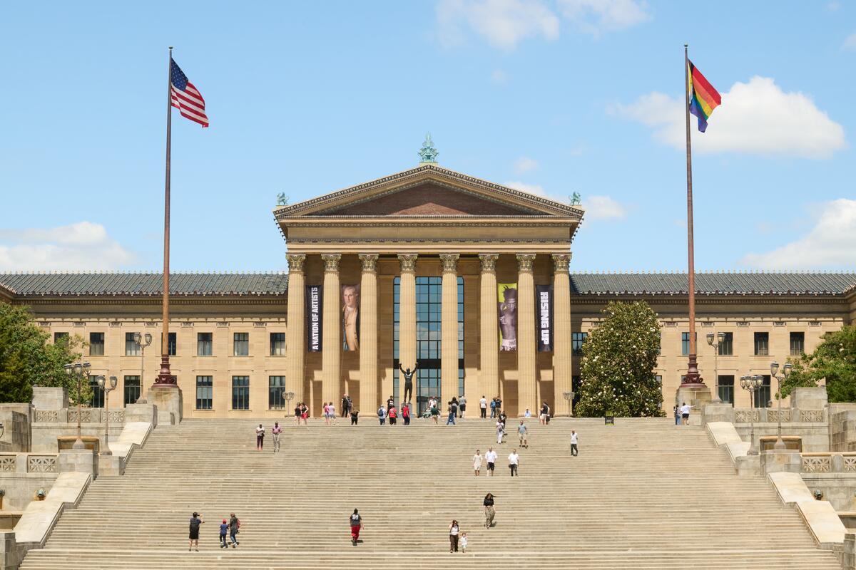 Beige neoclassical museum with tall columns, wide stairs, and US and Pride flags on flagpoles, with visitors on the steps.