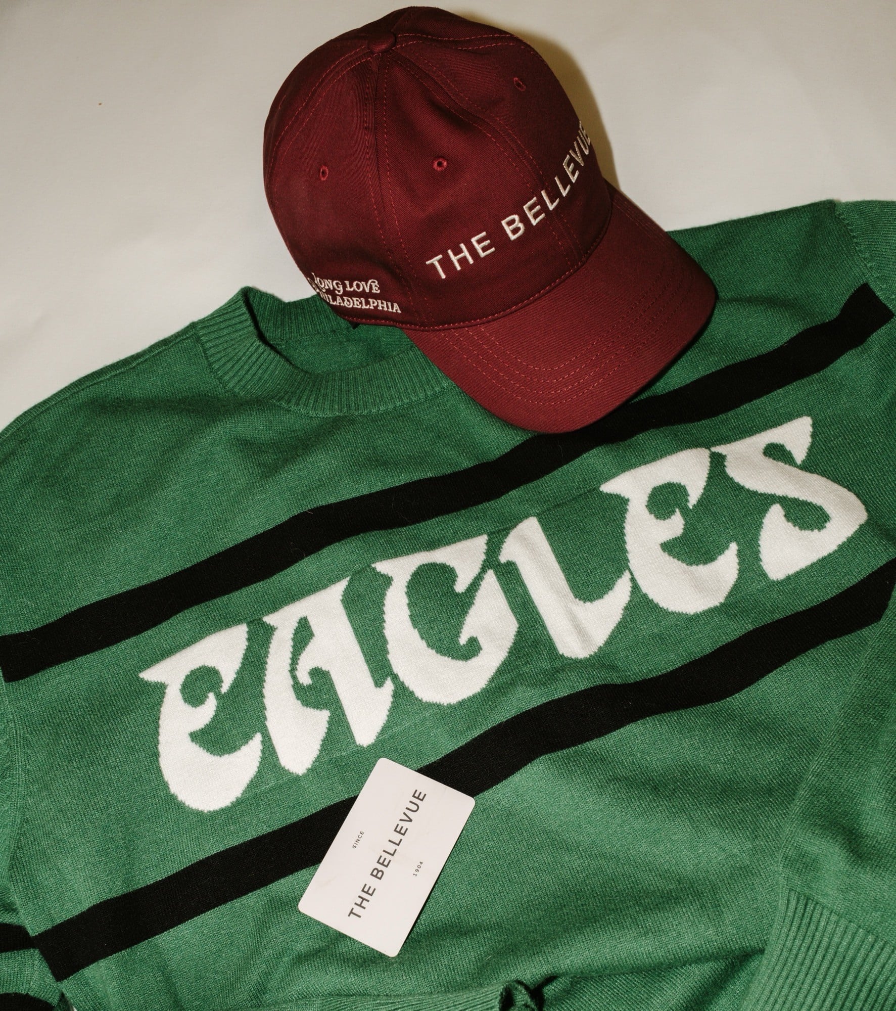 Maroon baseball cap with white embroidered text sits atop a green knit sweater with a large white graphic design visible underneath.