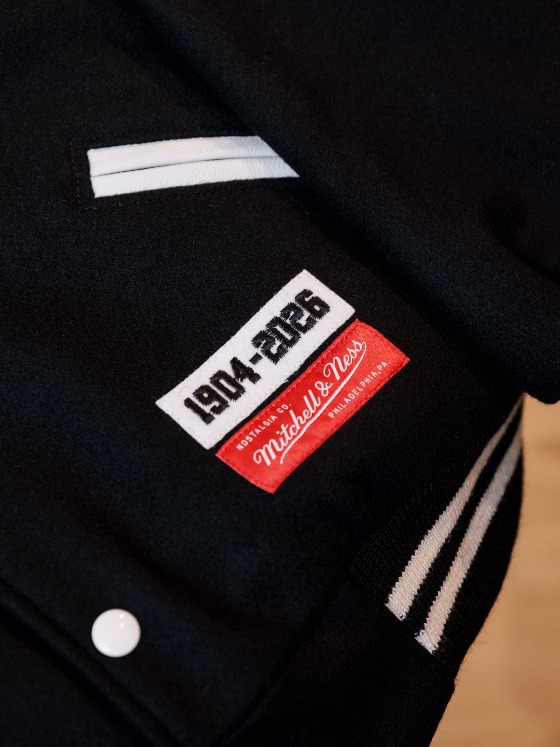 Close-up of a dark navy blazer showing a white stitched label pocket and a red Mitchell & Ness tag on the chest area.